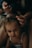 A young child with brown hair is sitting quietly while an adult gently styles their hair, using a small hair tie to create a hairstyle. The setting is intimate and dimly lit, giving a cozy and calm ambience. The child is wearing a white top and looks directly at the camera.