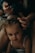 A young child with brown hair is sitting quietly while an adult gently styles their hair, using a small hair tie to create a hairstyle. The setting is intimate and dimly lit, giving a cozy and calm ambience. The child is wearing a white top and looks directly at the camera.