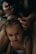A young child with brown hair is sitting quietly while an adult gently styles their hair, using a small hair tie to create a hairstyle. The setting is intimate and dimly lit, giving a cozy and calm ambience. The child is wearing a white top and looks directly at the camera.