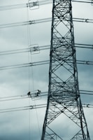 A tall metal electricity transmission tower stands against a cloudy sky. Two workers wearing safety helmets are suspended on a platform, performing maintenance work on the power lines.