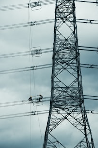 A tall metal electricity transmission tower stands against a cloudy sky. Two workers wearing safety helmets are suspended on a platform, performing maintenance work on the power lines.