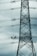 A tall metal electricity transmission tower stands against a cloudy sky. Two workers wearing safety helmets are suspended on a platform, performing maintenance work on the power lines.