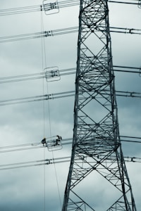 A tall metal electricity transmission tower stands against a cloudy sky. Two workers wearing safety helmets are suspended on a platform, performing maintenance work on the power lines.