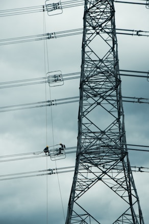 A tall metal electricity transmission tower stands against a cloudy sky. Two workers wearing safety helmets are suspended on a platform, performing maintenance work on the power lines.