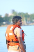 A person stands wearing a bright orange life jacket with a striped shirt underneath. Their hair appears wet, suggesting recent water activity. In the background, there is a blurred view of trees and possibly buildings, indicating an outdoor, waterside setting.