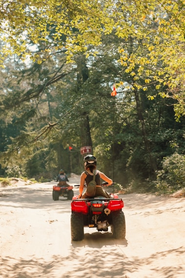 A person wearing a helmet rides an all-terrain vehicle (ATV) along a sandy path through a forested area, with another ATV visible ahead. The setting is lush with green trees and dappled sunlight breaking through the foliage.