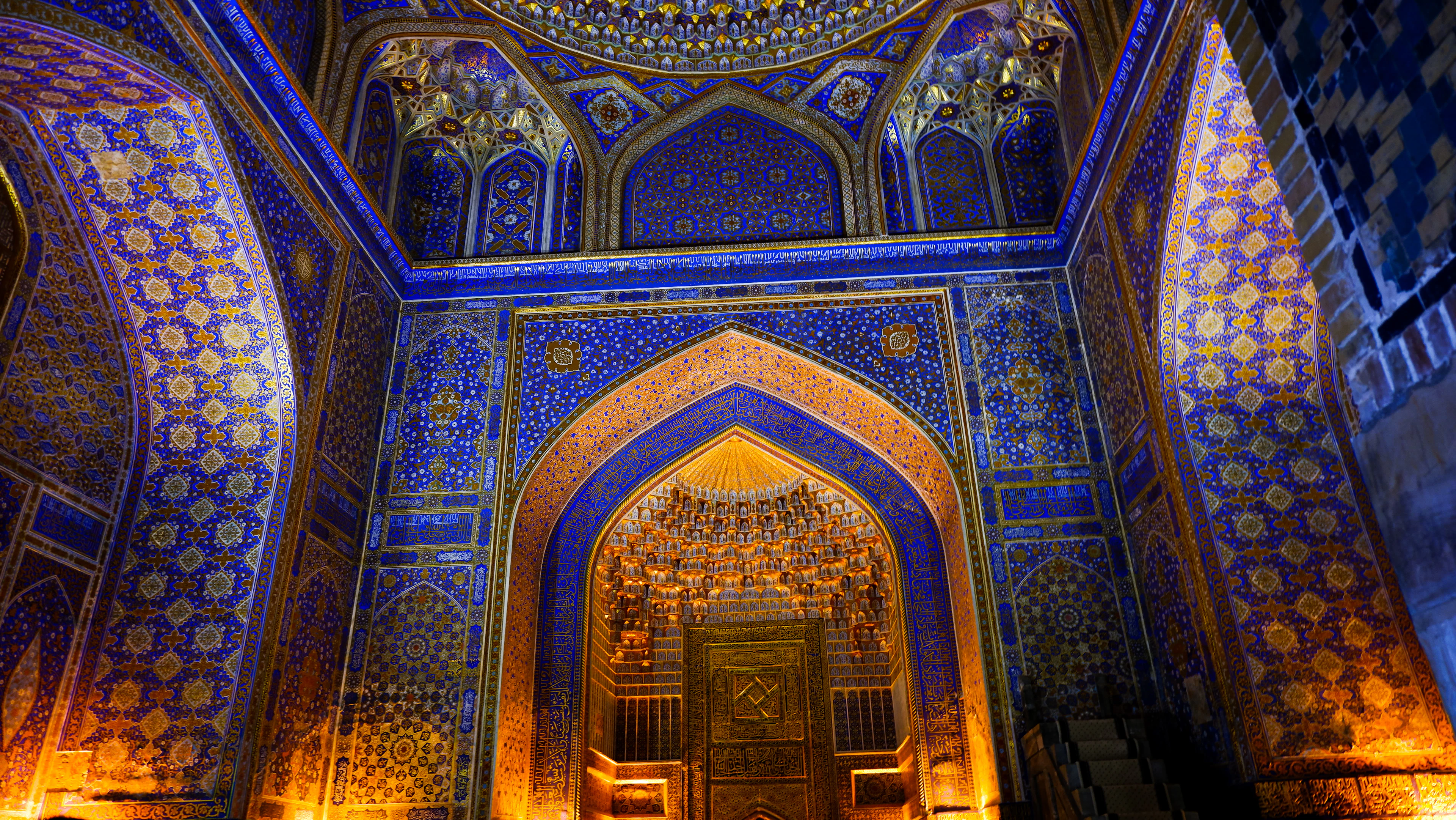 A gilded and blue-tiled prayer hall interior in Samarkand, its arched mihrab niche framed by dense geometric and floral patterning