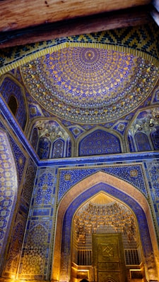 A beautifully intricate mosque interior features a richly decorated domed ceiling with geometric patterns and calligraphy. The walls are adorned with blue and gold motifs, creating a sense of opulence and spiritual reverence.