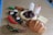 An assortment of plastic toy food items arranged on a wooden surface. Visible items include a loaf of bread, a slice of cheese, spaghetti with sauce, two baby bottles, sushi pieces, a red chili pepper, a green chili pepper, a bread roll, and a taco.