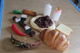 An assortment of plastic toy food items arranged on a wooden surface. Visible items include a loaf of bread, a slice of cheese, spaghetti with sauce, two baby bottles, sushi pieces, a red chili pepper, a green chili pepper, a bread roll, and a taco.