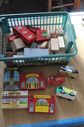 A collection of wooden toy blocks and vehicles is displayed on a wooden surface. A green plastic basket holds a mix of plain and decorated wooden blocks. In front of the basket, several wooden blocks resemble buildings, such as a town hall, school, grocery store, and fire station. Small wooden vehicles include a fire truck, a police car, and a bus.