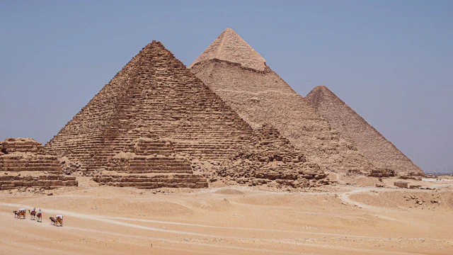 The three Giza pyramids rising from the desert plateau with tourists on camels passing in front under a clear blue sky.