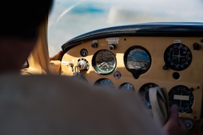A cockpit view with a close-up of aviation instruments, showcasing multiple dials and gauges. The lighting suggests natural daylight, and part of a person's hand is visible holding a control, indicating active operation of the aircraft.