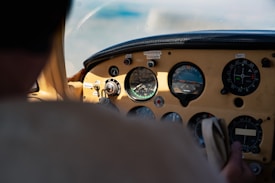 A cockpit view with a close-up of aviation instruments, showcasing multiple dials and gauges. The lighting suggests natural daylight, and part of a person's hand is visible holding a control, indicating active operation of the aircraft.