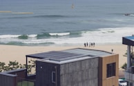 A seaside view with a modern building featuring a solar-paneled roof in the foreground. In the background, there is a sandy beach with waves approaching the shore. A small group of people stands close to the waterline, possibly looking out at the ocean. The sea appears calm and the sky is clear.