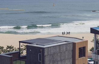 A seaside view with a modern building featuring a solar-paneled roof in the foreground. In the background, there is a sandy beach with waves approaching the shore. A small group of people stands close to the waterline, possibly looking out at the ocean. The sea appears calm and the sky is clear.