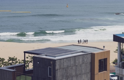 A seaside view with a modern building featuring a solar-paneled roof in the foreground. In the background, there is a sandy beach with waves approaching the shore. A small group of people stands close to the waterline, possibly looking out at the ocean. The sea appears calm and the sky is clear.