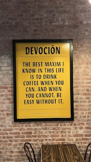A framed yellow poster with black text hangs on a brick wall. The poster features a quote by Jonathan Swift from 1772 about coffee. Below the poster, there is a wooden table with a curved metal chair visible.