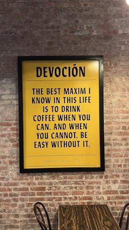 A framed yellow poster with black text hangs on a brick wall. The poster features a quote by Jonathan Swift from 1772 about coffee. Below the poster, there is a wooden table with a curved metal chair visible.