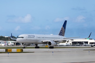 A passenger airplane is on a runway at an airport. The aircraft has 'United' written on its side and features a dark blue tail with a globe emblem. Several other airplanes and airport buildings are visible in the background under a partly cloudy sky.