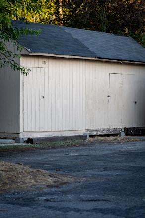A simple wooden shed with a pitched roof, painted in a light cream color. The shed has two doors and is situated on a rough, cracked asphalt ground. Trees with green foliage are visible in the background on the left side of the image.