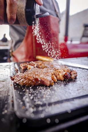 A close-up of a person seasoning meat with salt on a hot grill. The salt is being sprinkled from a shaker held in the hand above the grilled meat, which appears to be steak. The focus is on the falling salt and meat, with the background slightly blurred, showing someone wearing a red apron.