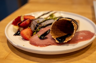 A white plate with a dessert that includes a waffle cone, slices of banana and strawberries, ice cream or sorbet, and chocolate sauce drizzled on top. The ice cream or sorbet is melting, creating an inviting and appetising display. The plate is set on a wooden surface.
