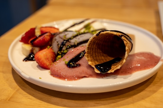 A white plate with a dessert that includes a waffle cone, slices of banana and strawberries, ice cream or sorbet, and chocolate sauce drizzled on top. The ice cream or sorbet is melting, creating an inviting and appetising display. The plate is set on a wooden surface.