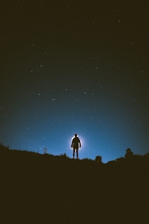 Silhouetted figure standing atop a hill against a deep bottle green sky with an eerie glow