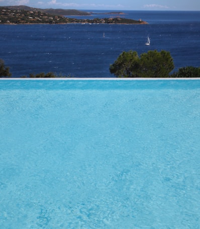 A serene infinity pool overlooks a vast expanse of deep blue ocean, with small sailboats dotting the distant waters. Behind the pool, there are lush green trees and a coastline lined with hills and scattered houses. The sky is clear with a few clouds on the horizon, adding to the tranquil atmosphere.