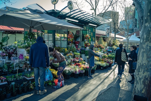 A lively outdoor flower market with colorful arrangements displayed on stands. Several people are browsing and purchasing flowers, with one person handing over a bouquet. The market is shaded by a large umbrella, and the setting is urban with buildings in the background.