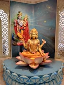 A detailed sculpture depicting Hindu deities, with one central figure sitting on a lotus flower displaying multiple arms, adorned with traditional ornaments and a serene expression. Behind, a backdrop featuring a cosmic theme with stars and galaxies enhances the divine atmosphere. Two additional figures, also richly dressed and posed, are positioned in the background alongside a peacock.