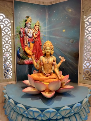 A detailed sculpture depicting Hindu deities, with one central figure sitting on a lotus flower displaying multiple arms, adorned with traditional ornaments and a serene expression. Behind, a backdrop featuring a cosmic theme with stars and galaxies enhances the divine atmosphere. Two additional figures, also richly dressed and posed, are positioned in the background alongside a peacock.