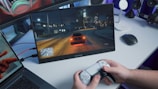 A person is holding a gaming controller in front of a screen showing a nighttime driving scene in a video game. The screen is placed on a desk alongside a laptop and a wireless mouse. The room has a blue ambient lighting.