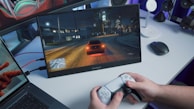 A person is holding a gaming controller in front of a screen showing a nighttime driving scene in a video game. The screen is placed on a desk alongside a laptop and a wireless mouse. The room has a blue ambient lighting.