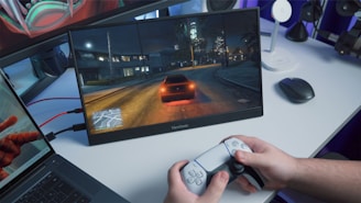 A person is holding a gaming controller in front of a screen showing a nighttime driving scene in a video game. The screen is placed on a desk alongside a laptop and a wireless mouse. The room has a blue ambient lighting.