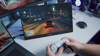 A person is holding a gaming controller in front of a screen showing a nighttime driving scene in a video game. The screen is placed on a desk alongside a laptop and a wireless mouse. The room has a blue ambient lighting.
