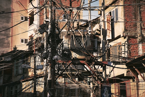 A dense network of overhead wires is tangled around utility poles in front of an aged, multistory building with exposed brick and decaying walls. Some air conditioning units are mounted on the walls, and various windows are visible. The chaotic arrangement of cables suggests a possibly dangerous or deteriorating infrastructure.