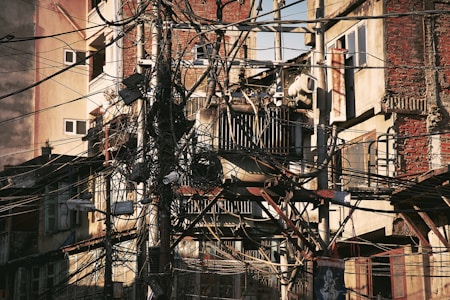 A dense network of overhead wires is tangled around utility poles in front of an aged, multistory building with exposed brick and decaying walls. Some air conditioning units are mounted on the walls, and various windows are visible. The chaotic arrangement of cables suggests a possibly dangerous or deteriorating infrastructure.