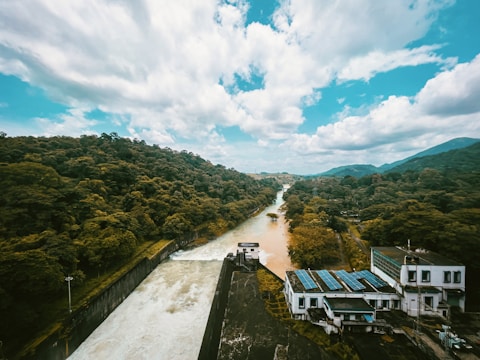 A scenic view of a river cutting through a lush green forest under a partly cloudy sky. On the right, a hydroelectric facility is visible with solar panels on its roof. The river flows smoothly between dense vegetation, with some light reflecting off the water's surface.