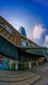 A modern urban architecture scene featuring a tall skyscraper with reflective blue glass windows against a clear blue sky. The building's facade includes a multi-story glass atrium, with geometric patterns and digital advertisements. The pavement in front of the building features a circular, striped design leading towards the entrance.