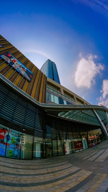 A modern urban architecture scene featuring a tall skyscraper with reflective blue glass windows against a clear blue sky. The building's facade includes a multi-story glass atrium, with geometric patterns and digital advertisements. The pavement in front of the building features a circular, striped design leading towards the entrance.
