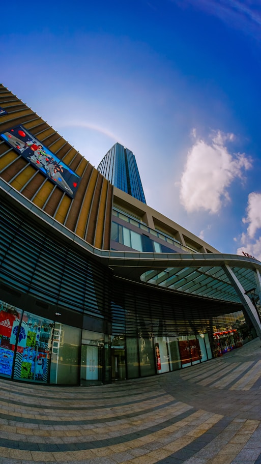 A modern urban architecture scene featuring a tall skyscraper with reflective blue glass windows against a clear blue sky. The building's facade includes a multi-story glass atrium, with geometric patterns and digital advertisements. The pavement in front of the building features a circular, striped design leading towards the entrance.