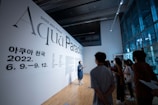 A group of people in a modern, well-lit gallery space are gathered around listening to a person giving a presentation. The wall features large text reading 'Aqua Paradiso' and includes dates in 2022, likely indicating the title and timeline of an exhibition or event. The room has large windows and sleek architecture, creating an open and contemporary environment.