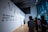 A group of people in a modern, well-lit gallery space are gathered around listening to a person giving a presentation. The wall features large text reading 'Aqua Paradiso' and includes dates in 2022, likely indicating the title and timeline of an exhibition or event. The room has large windows and sleek architecture, creating an open and contemporary environment.