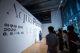 A group of people in a modern, well-lit gallery space are gathered around listening to a person giving a presentation. The wall features large text reading 'Aqua Paradiso' and includes dates in 2022, likely indicating the title and timeline of an exhibition or event. The room has large windows and sleek architecture, creating an open and contemporary environment.