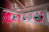 The image features a modern art gallery with a striking mural on the walls. The mural consists of several large, black and white geometric spheres with intricate patterns, set against a vivid pink background with dynamic white streaks. The floor is wooden, and the ceiling has a contemporary design with spotlights.
