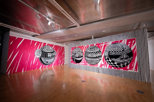 The image features a modern art gallery with a striking mural on the walls. The mural consists of several large, black and white geometric spheres with intricate patterns, set against a vivid pink background with dynamic white streaks. The floor is wooden, and the ceiling has a contemporary design with spotlights.