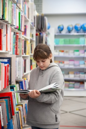 A person wearing a gray hoodie is standing in a library aisle, reading a book. Shelves filled with colorful books line the aisle. In the background, globes are visible, adding an educational atmosphere.