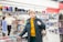 A person wearing a dark green jacket and yellow turtleneck is standing in a store with shelves filled with books and stationery items. Another person in a beige jacket is browsing through the shelves in the background.